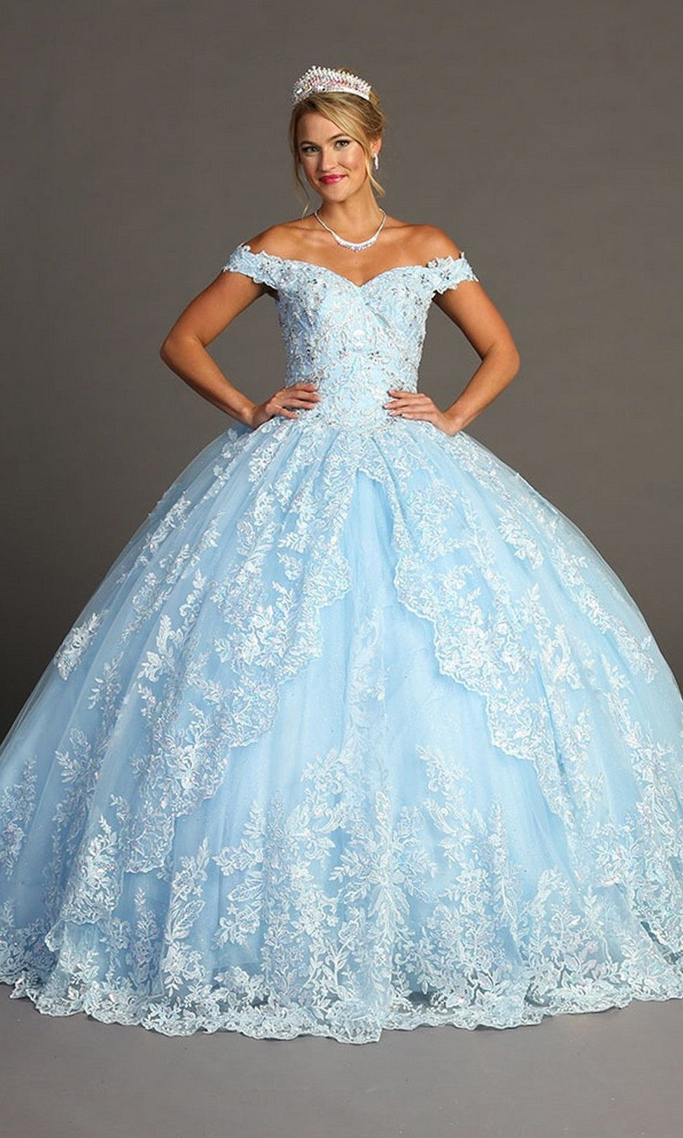 A woman wearing the Quinceanera Dress CHF3072 By Chicas in sky blue, featuring an off-the-shoulder design with delicate lace details and touches of metallic embroidery, accessorized with a tiara and necklace, stands against a plain background.
